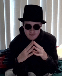 a man wearing a black hat and sunglasses is sitting in front of a window