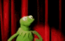a green kermit the frog is standing in front of a red curtain .