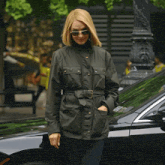 a woman wearing sunglasses and a green jacket is standing in front of a black car