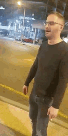a man wearing glasses and a black shirt is standing on the side of a street .