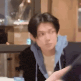 a man in a black jacket and blue hoodie is sitting at a table looking at a tablet .