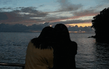 two women are hugging each other in front of a body of water at sunset .