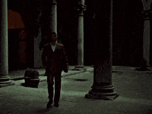 a man in a brown suit and white shirt stands in a dark courtyard