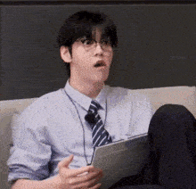 a young man wearing glasses and a tie is sitting on a couch holding a tablet computer .
