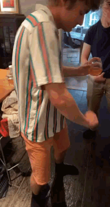 a young man in a striped shirt and orange shorts is standing in a room holding a glass of beer .