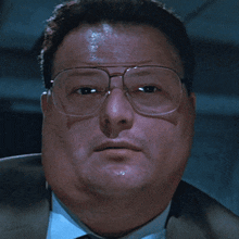 a man wearing glasses and a suit looks at the camera with a serious look on his face