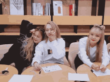 three girls are sitting at a table with one wearing a white sweatshirt that says ' nct ' on it