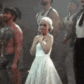 a woman in a white dress is standing in front of a group of men in white tank tops