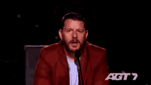 a man in a red suit is speaking into a microphone with the letters agt 7 behind him