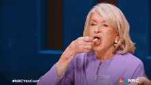 a woman in a purple jacket is eating something with the nbc logo behind her