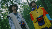 a man in a hat and a man in a colorful jacket are standing in the woods