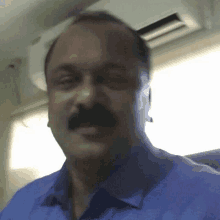 a man with a mustache is wearing a blue shirt and sitting in front of a window