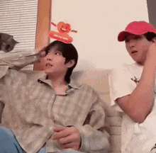 two men are sitting on a couch with a cat . one of the men is wearing a pumpkin hat on his head .