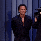 a man in a suit stands in front of a blue curtain while a man holds a camera