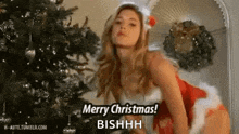 a woman in a santa claus outfit is standing in front of a christmas tree and saying merry christmas .