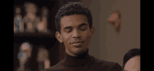 a man in a brown turtleneck sweater is smiling in front of a shelf full of statues .