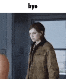 a woman in a brown jacket is standing in front of a window and says bye