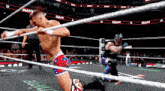 a wrestler is kneeling down in a wrestling ring while another wrestler is standing in the ropes .