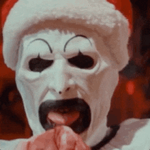 a close up of a clown wearing a santa hat and a mask with his tongue out .