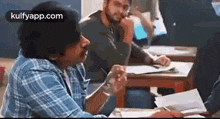 a man in a plaid shirt is sitting at a desk in a classroom talking to another man .
