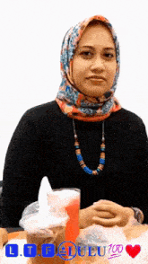 a woman wearing a scarf and a necklace sits at a table with a cup of red liquid