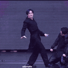 a man in a black suit is dancing on a stage while another man kneeling down behind him .