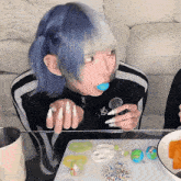 a girl with blue hair and white nails is sitting at a table eating candy