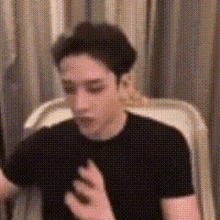 a man in a black shirt is sitting in a chair with his hand on his chest .