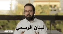 a man with a beard and glasses is sitting at a table and talking in a foreign language .
