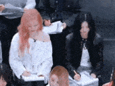 a woman with pink hair is sitting in a crowd of people writing in notebooks