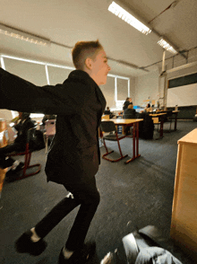 a boy in a black suit is jumping in the air in a classroom
