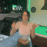 a woman stands in front of a pool table with a dart board on the wall above her