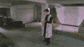 a man in a white coat stands in an empty room with boxes and a ladder