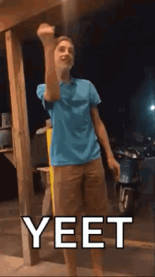 a young man in a blue shirt and brown shorts is standing in front of a scooter and making a fist .