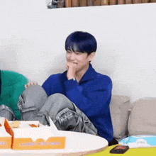 a man with blue hair is sitting on a couch with his legs crossed and eating a donut .