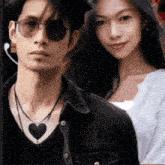 a man wearing sunglasses and a necklace with a heart on it stands next to a woman