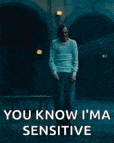 a man in a white sweater is standing in a dark room with the words `` you know i 'm sensitive '' .