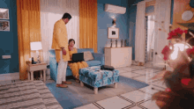 a man in a yellow jacket stands next to a woman sitting on a blue couch using a laptop