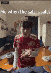a man wearing a red shirt that says " me when the salt is salty " is standing in a messy room