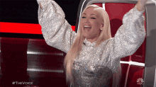 a woman in a silver dress is sitting in a chair with her arms in the air and the words #thevoice above her