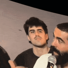 a man with a beard is talking into a microphone while another man watches .