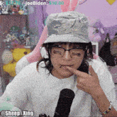 a woman wearing a hat and glasses is eating something in front of a microphone with the hashtag @sheep_xing visible