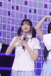a girl in a white shirt and blue skirt is holding a microphone and dancing on a stage .