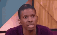 a man in a purple shirt is sitting in front of a wooden wall and making a funny face .