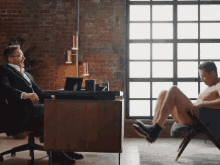 a man in a suit sits at a desk next to a man without a shirt on