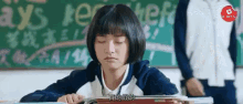 a girl is sitting at a desk in a classroom with her eyes closed and a man standing behind her .