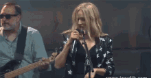 a woman singing into a microphone with a man playing a guitar behind her .