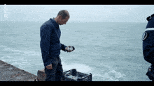 a man in a blue jacket stands on a ledge overlooking the ocean with a badge that says police