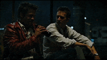 a man in a red leather jacket sits next to another man in a white shirt smoking a cigarette