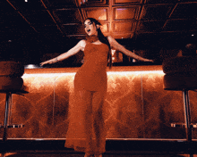a woman in a red dress stands in front of a bar with her arms outstretched